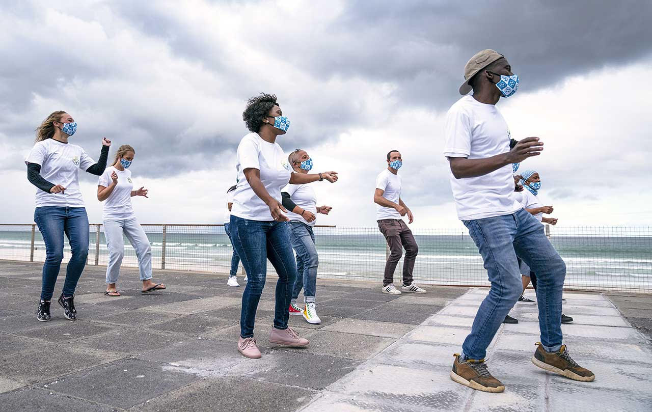 A group of young people of mixed race and gender and wearing jeans, white t-shirts and protective face masks perform a dance next to a beach.